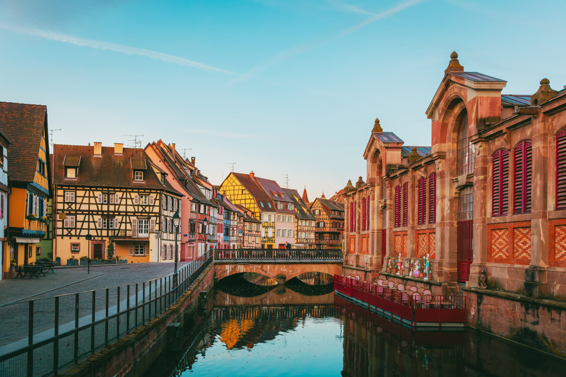 The red-brick Covered Market stands beside a canal in Colmar, France, facing colourful half-timbered buildings and a small stone bridge.