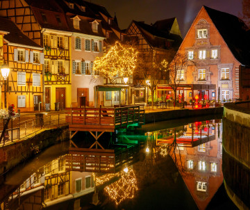 Night view of Little Venice in Colmar, France, with illuminated half-timbered houses, a decorated tree and historic buildings reflected in the canal.