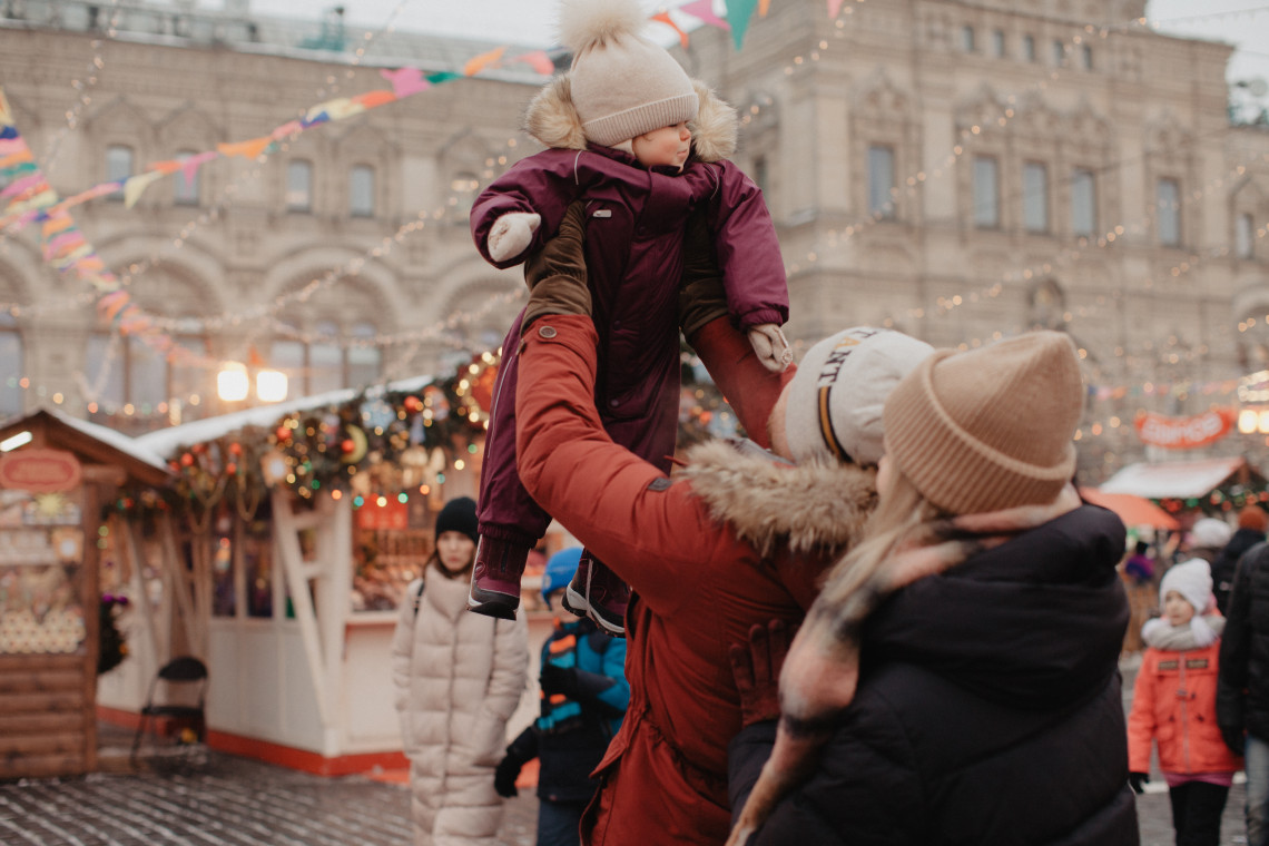A family with a young child visits the Christmas market in Red Square, Moscow, Russia. Decorated wooden stalls, seasonal lights and the historic GUM department store appear in the background.