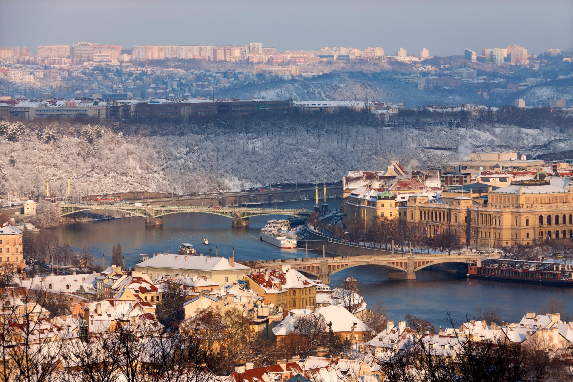 Winter panorama of the Vltava River in Prague, Czech Republic, with snow-covered rooftops, wooded hills and bridges spanning the water. Riverboats and historic buildings extend along both banks.
