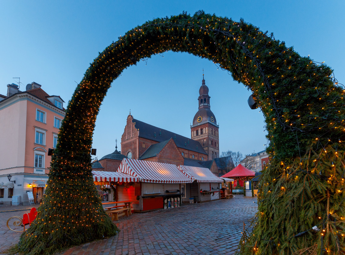 An illuminated evergreen arch frames the Christmas market beside Riga Cathedral in Riga, Latvia. Red-and-white market stalls stand across the cobbled square beneath the cathedral&rsquo;s clock tower.