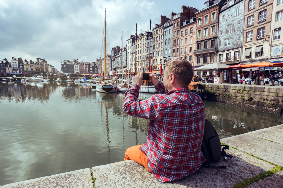 Photographer sitting on a pier capturing Honfleur's colorful harbor with tall Norman houses, Normandy, France