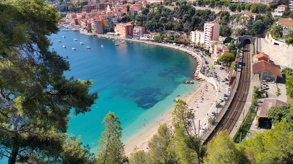 Aerial view of Villefranche-sur-Mer turquoise bay with sandy beach, colorful buildings and railway line along the French Riviera, France