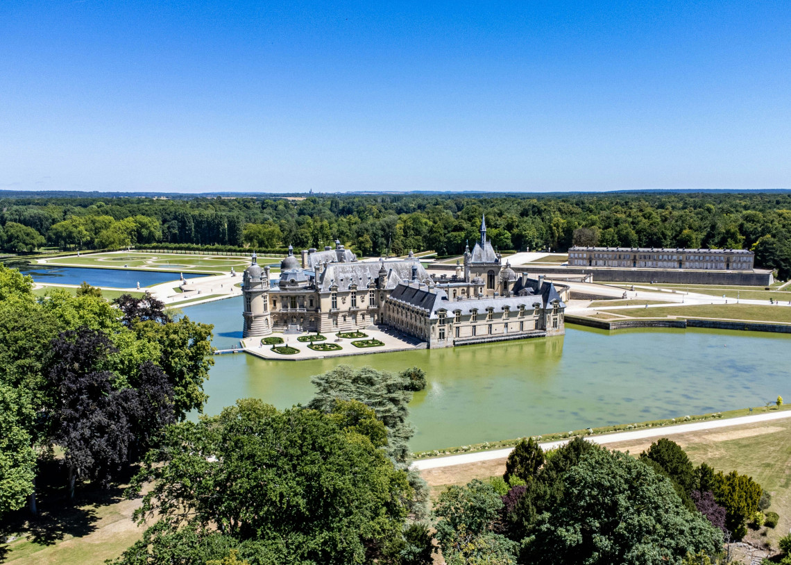 Aerial view of Chateau de Chantilly surrounded by a wide moat with formal French gardens and forested parkland stretching into the distance in northern France