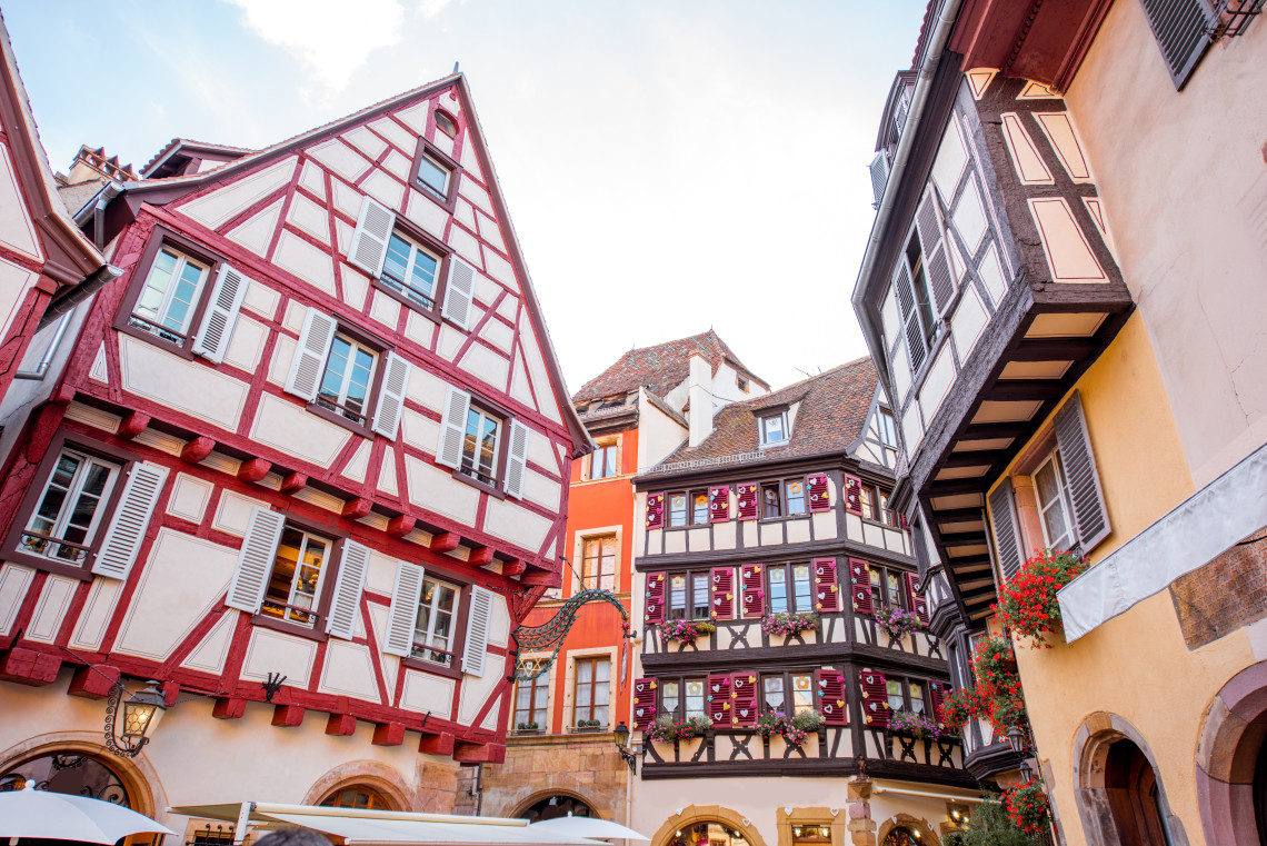 Close-up view of half-timbered Alsatian buildings in Colmar's old town, with bold red and white timber framing, overhanging upper storeys, wooden shutters, flower boxes, and a street lantern at the corner