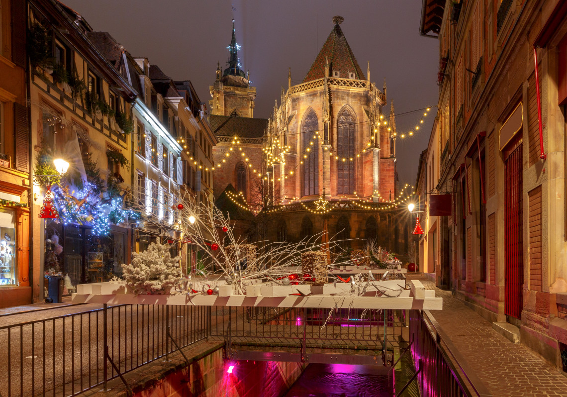  Kaysersberg at night during the Christmas market season, with a decorated bridge over the river in the foreground, string lights and Christmas decorations lining the street, and the illuminated sandstone Gothic church tower glowing in the background