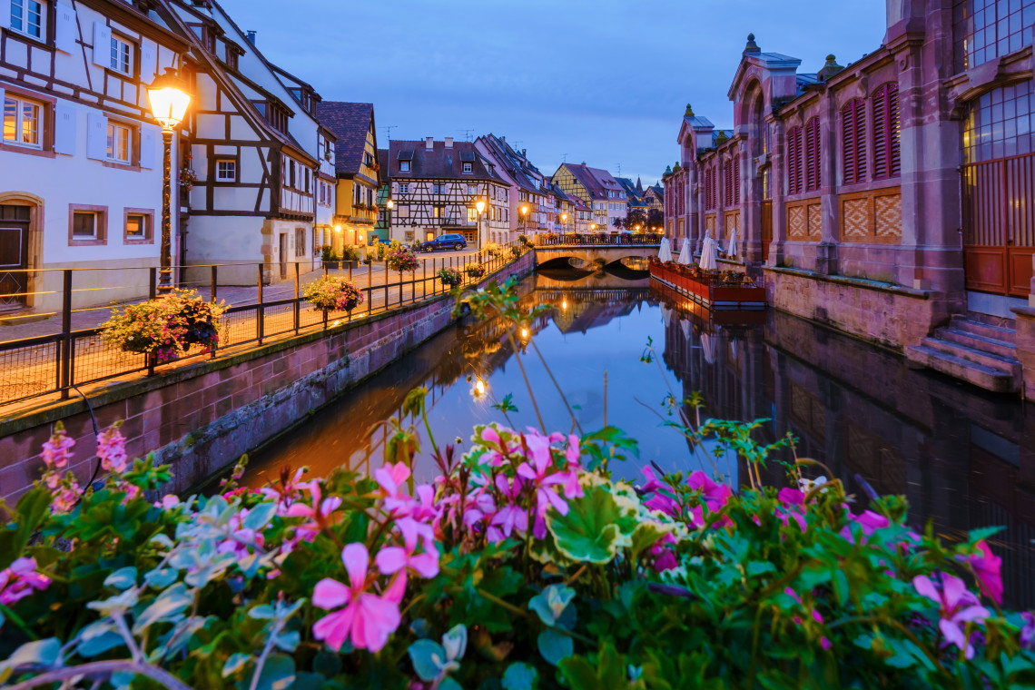 Dusk view of the Lauch canal in Colmar with pink geraniums in sharp focus in the foreground, warm street lamp reflections in the still water, half-timbered buildings lining the left bank, and the pink sandstone facade of the Marché Couvert on the right under a blue evening sky