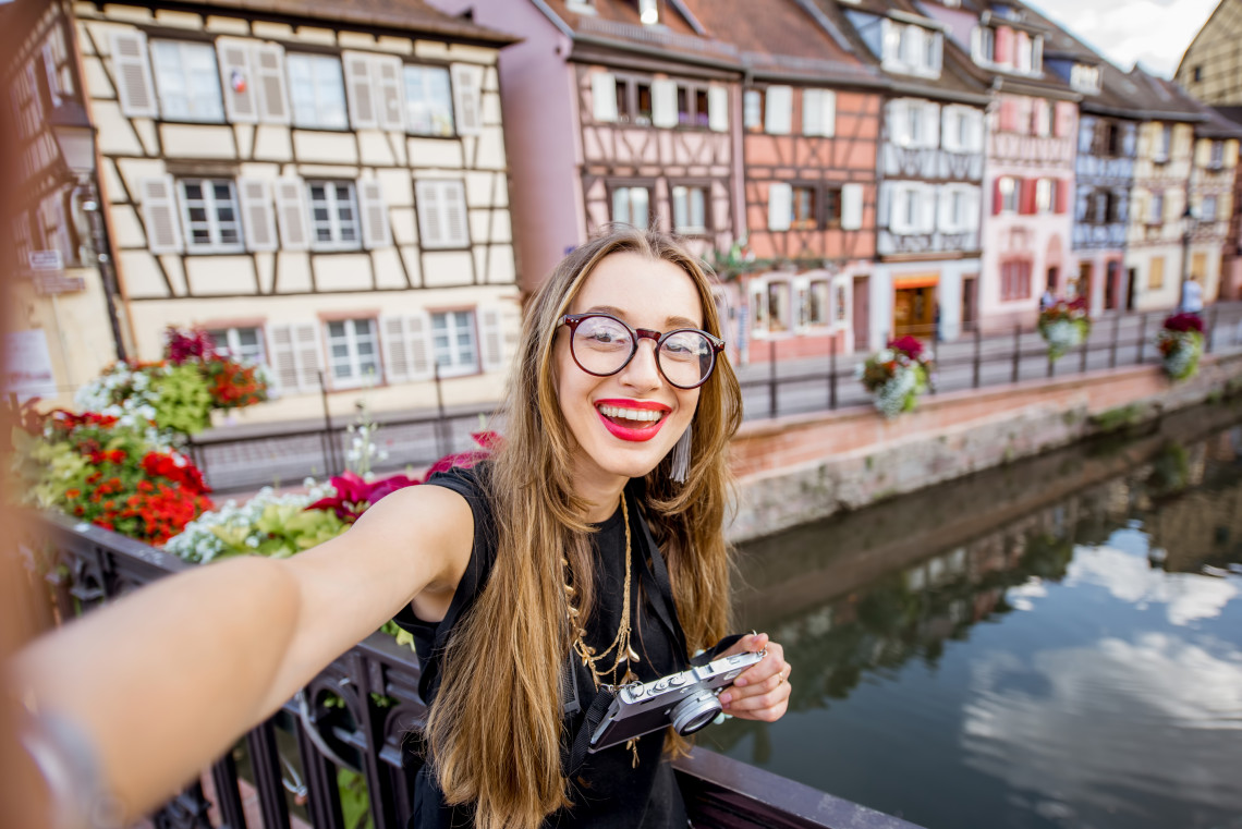 A smiling young woman with glasses and a film camera takes a selfie on a canal bridge in Colmar's Little Venice quarter, with colourful half-timbered Alsatian buildings and red geraniums blurred in the background
