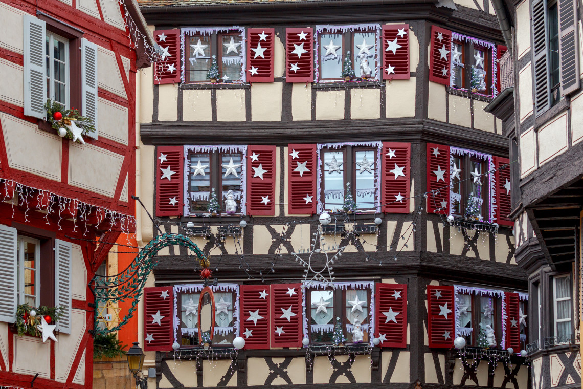Close-up of a half-timbered Colmar building during the Christmas market season, with red shutters, white star decorations on every window, icicle lights, and Christmas wreaths on the facade