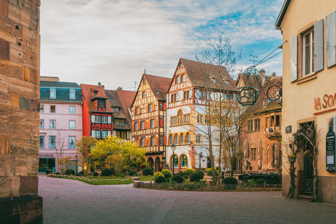 A quiet cobblestone square in Colmar with a mix of half-timbered and painted Alsatian buildings, early spring trees in a central garden, a wine bar sign on the right, and a medieval stone wall in the foreground