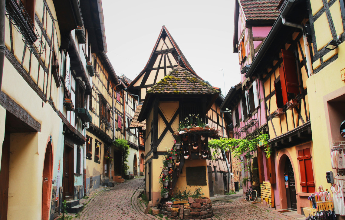 The cobblestone Place du Puits in Eguisheim on the Alsace Wine Route, with a central half-timbered well structure topped by a moss-covered pointed roof, colourful half-timbered buildings in yellow, pink and ochre lining the narrow street on both sides, and hanging flower baskets above small shops