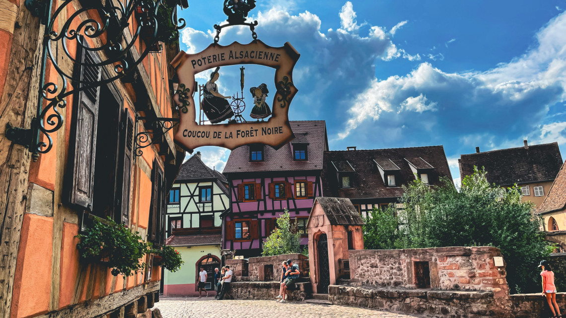  An ornate wrought-iron shop sign for a Poterie Alsacienne framing a view of colourful half-timbered buildings, a medieval stone wall with a small gatehouse chapel, and visitors sitting in the sun near the Dolder gate in Riquewihr