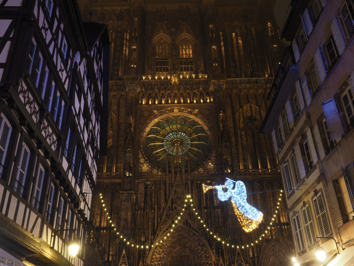 Strasbourg Cathedral rises above a narrow street in Strasbourg, France, with its Gothic fa&ccedil;ade and stained-glass rose window illuminated at night. Strings of lights and a glowing angel decoration mark the Christmas season.