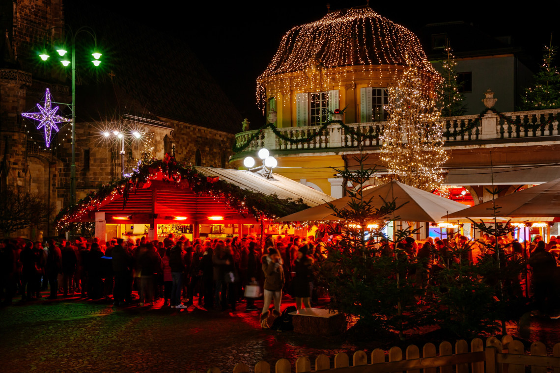 Visitors gather around illuminated wooden stalls at the Christmas market in Baden-Baden, Germany. Warm lights cover the nearby pavilion and seasonal decorations across the busy market square.