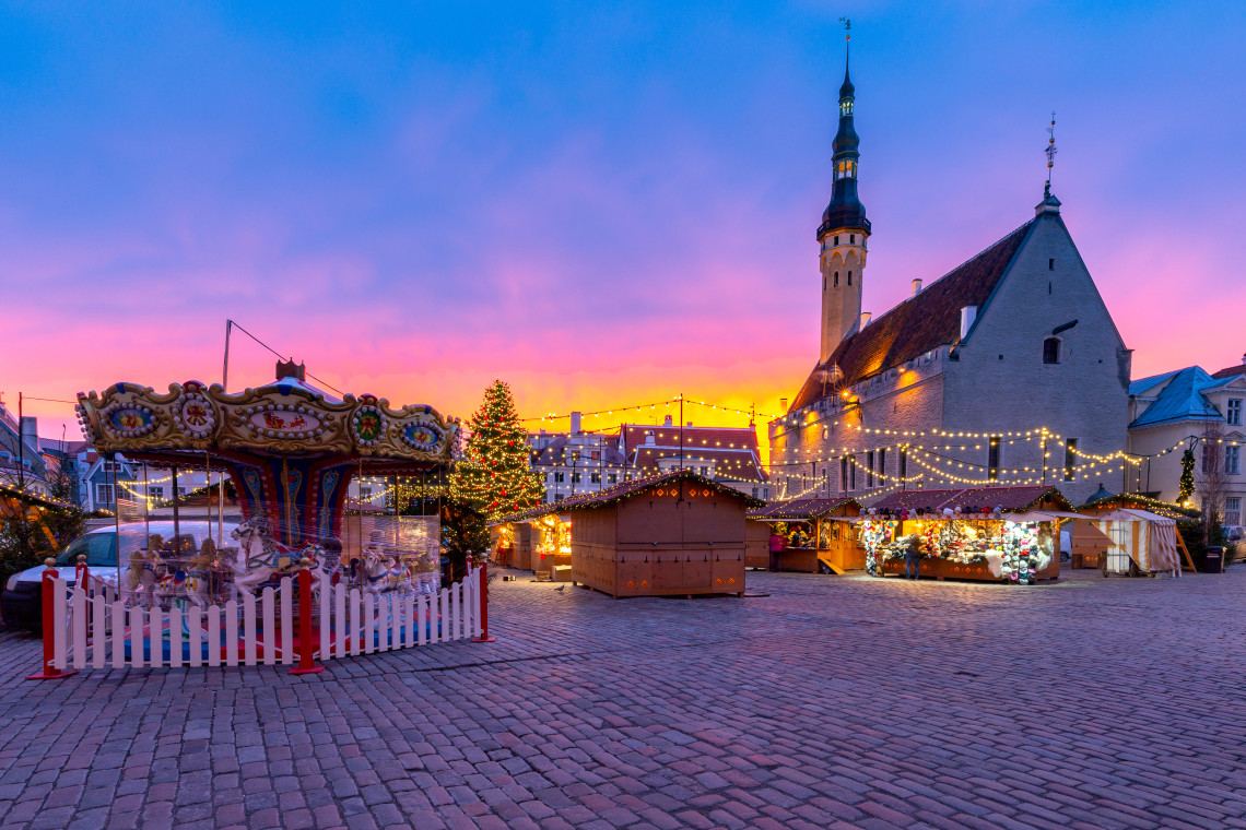 A traditional carousel, decorated Christmas tree and illuminated wooden stalls fill Tallinn Christmas Market in Town Hall Square, Tallinn, Estonia. The medieval Tallinn Town Hall rises behind the cobbled square at sunset.