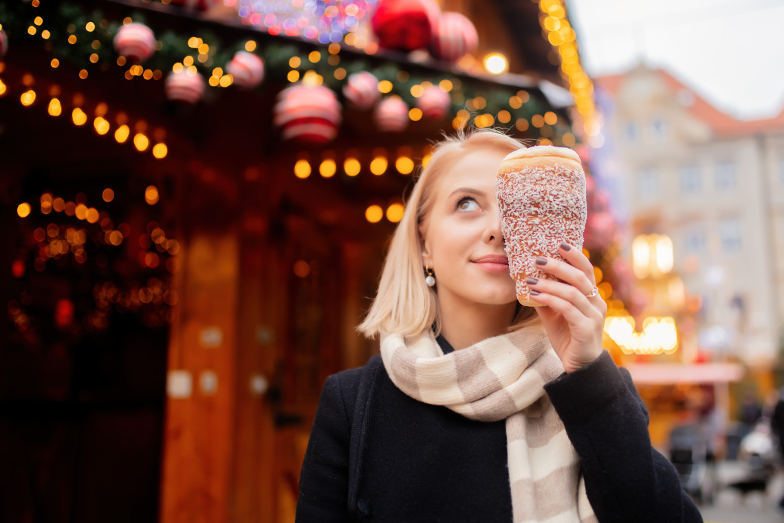 A visitor holds a freshly prepared chimney cake coated with sugar at a Christmas market in Budapest, Hungary. Illuminated wooden stalls and hanging ornaments create a softly blurred festive background.