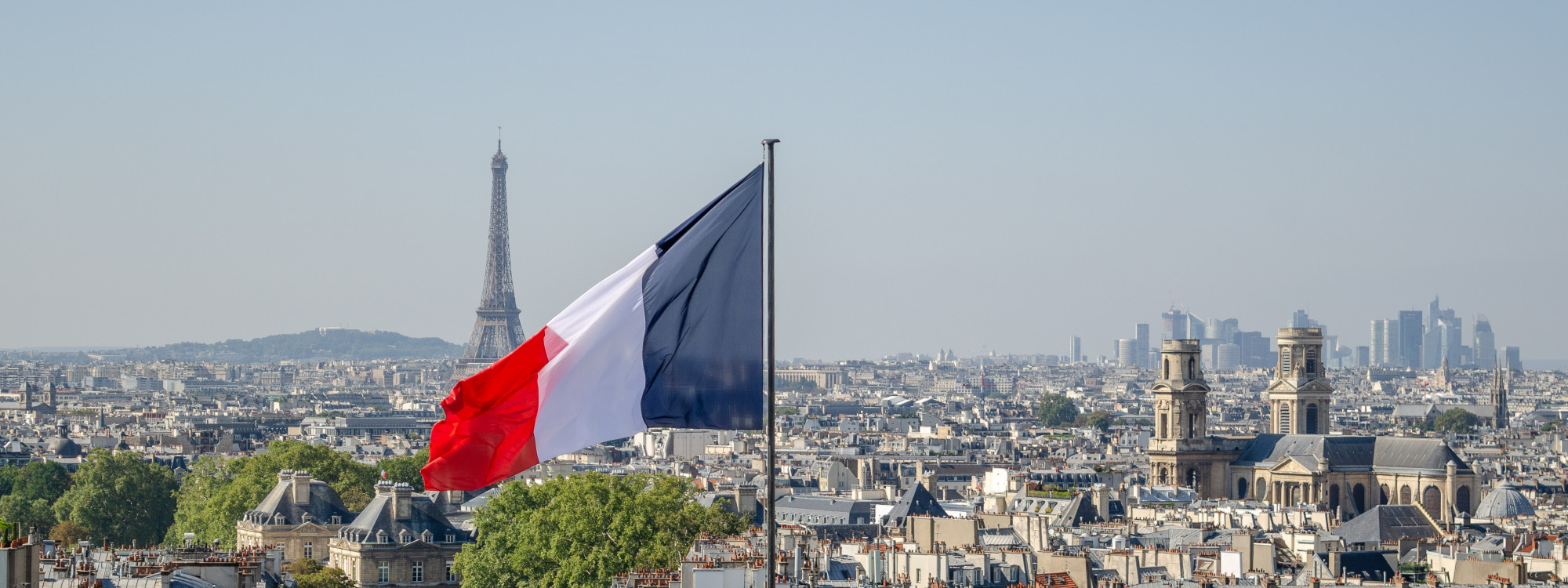 French flag flying over the Paris skyline with the Eiffel Tower and La Défense business district in the background, France