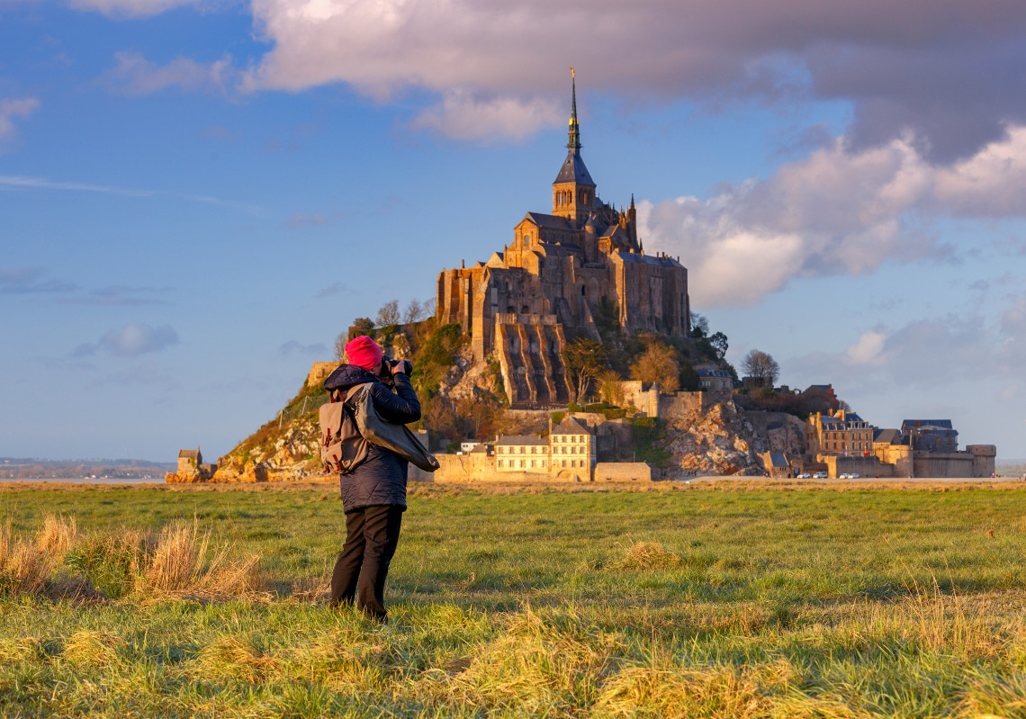 Photographer in warm winter clothes photographing Mont Saint-Michel abbey from the salt marshes in Normandy, France