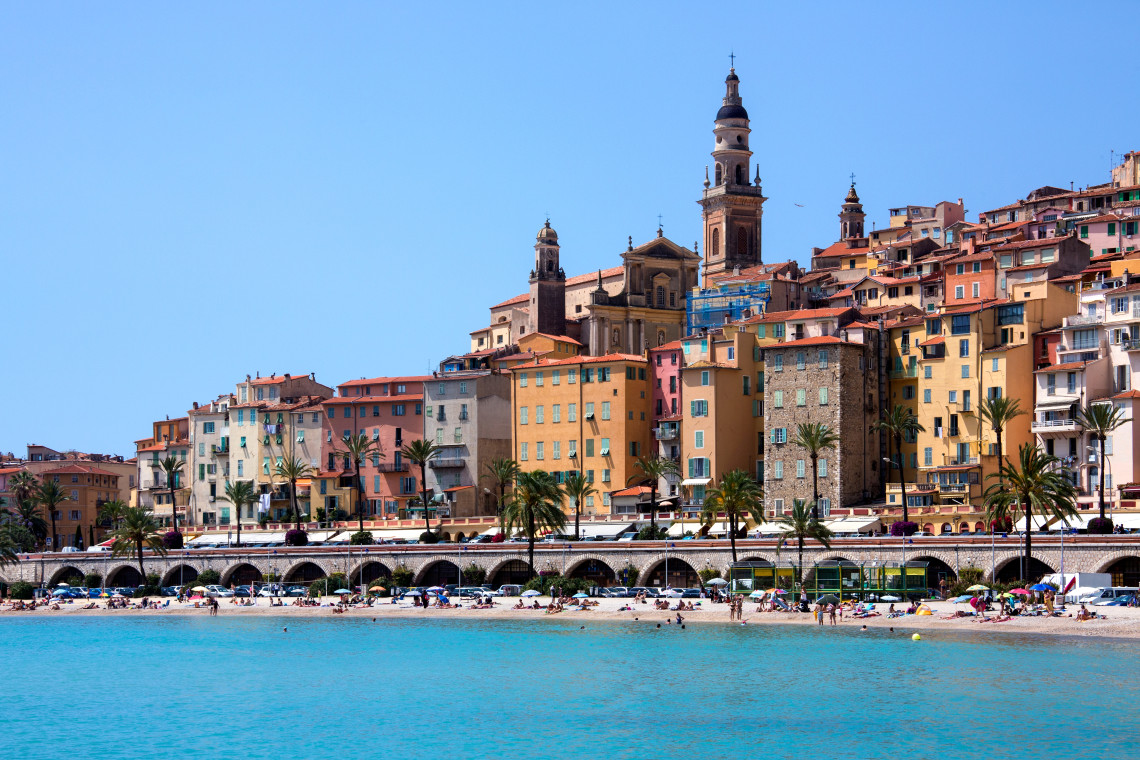 Colorful buildings and church towers of Menton old town line the waterfront above a sandy beach on the French Riviera