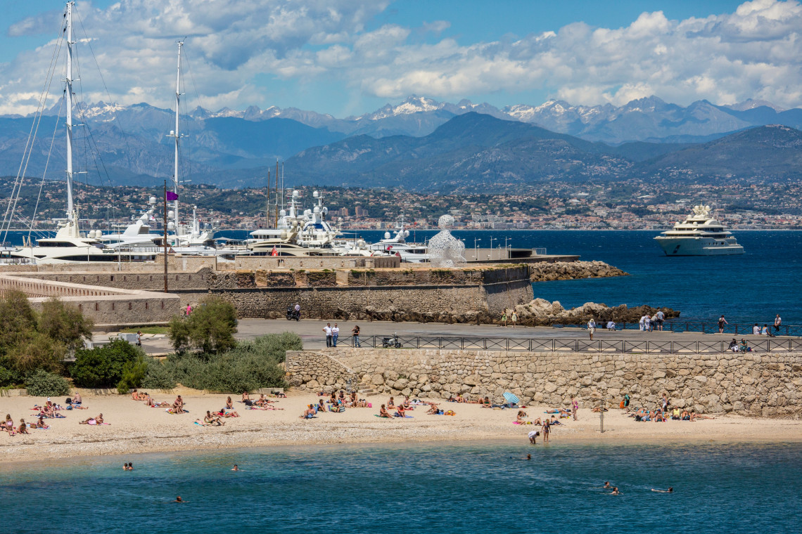 Sunbathers on the beach in Antibes with the marina packed with yachts and the snow capped Alps visible in the background on the French Riviera