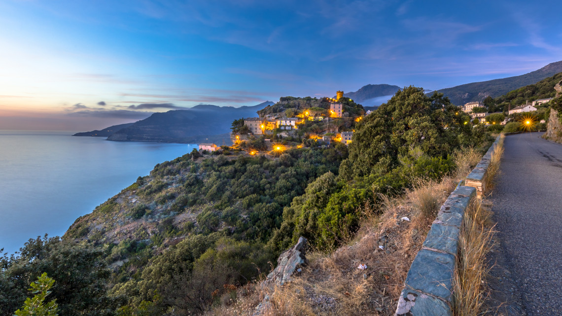 A hilltop village lit up at dusk on a rocky promontory overlooking the sea and mountains in Corsica France
