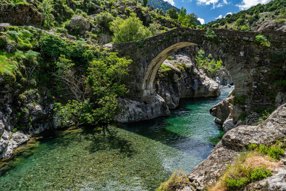 A historic single arch stone bridge spanning a clear green river cutting through a rocky gorge surrounded by lush vegetation in Corsica France