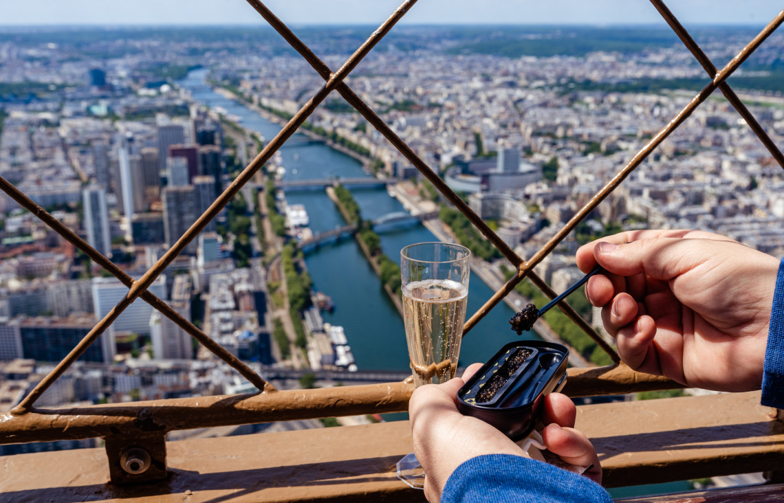 Hands holding a champagne flute and caviar on the top floor of the Eiffel Tower with the Seine River and Paris skyline below, France