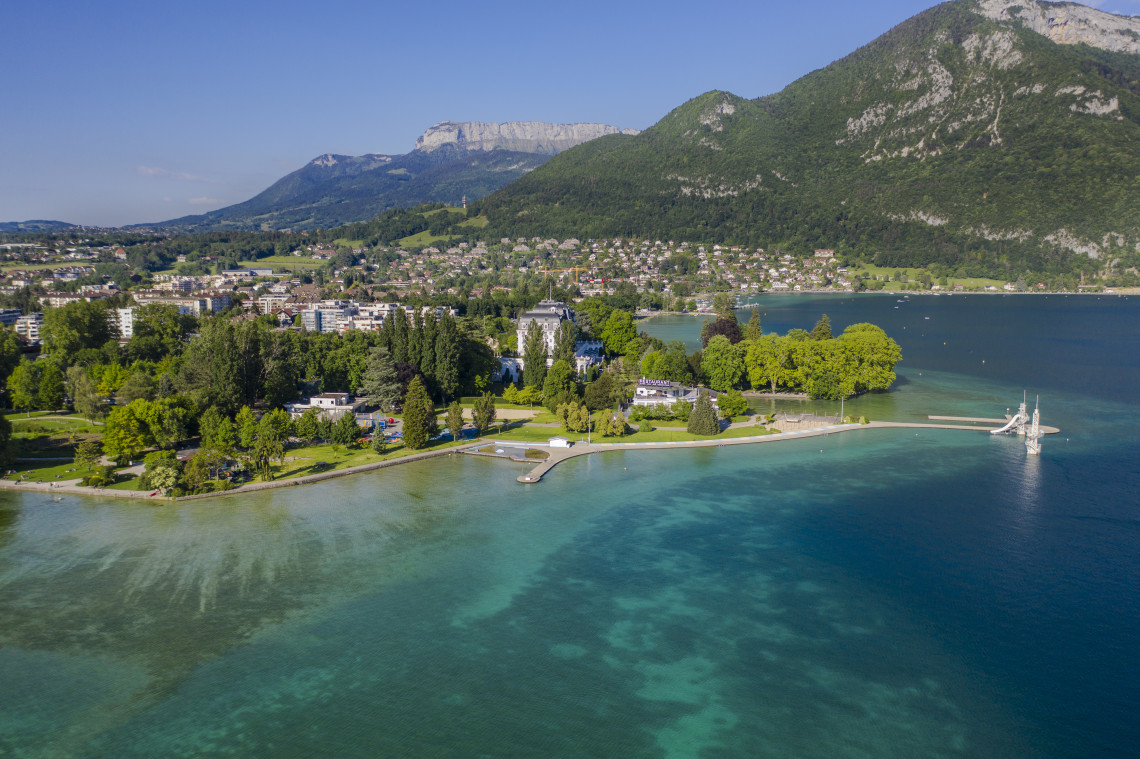 Aerial view of the Annecy lakeside showing turquoise water with visible underwater clarity, a tree-lined promenade, a white Belle &Eacute;poque villa, an ornate iron jetty extending into the lake, and limestone Alpine ridges rising behind the town