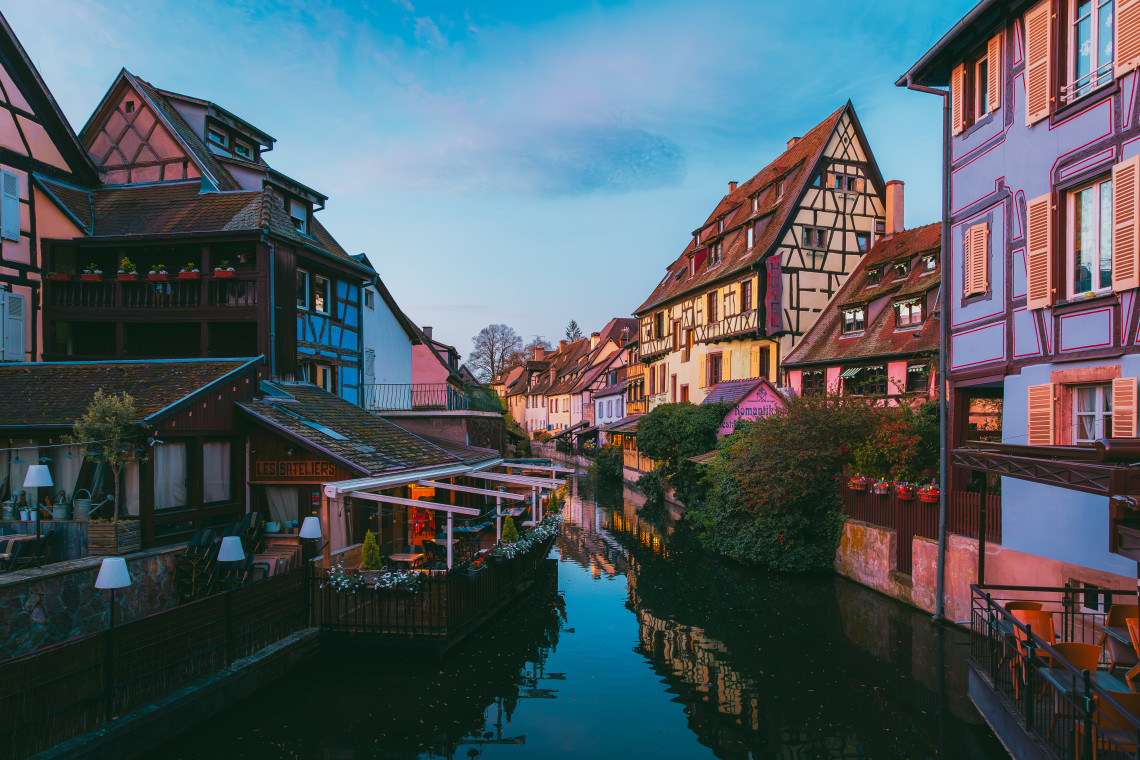 Blue hour view along the Lauch canal in Colmar's Little Venice quarter, with colourful half-timbered Alsatian buildings in pink, yellow and orange reflected in the still water, a restaurant terrace with warm lighting on the left, and flower boxes on the facades