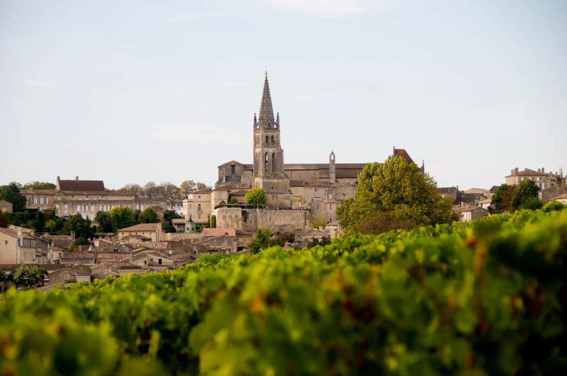 The medieval hilltop village of Saint-&Eacute;milion in Bordeaux wine country, with the spire of the Monolithic Church rising above limestone rooftops, framed by blurred green vineyard rows in the foreground
