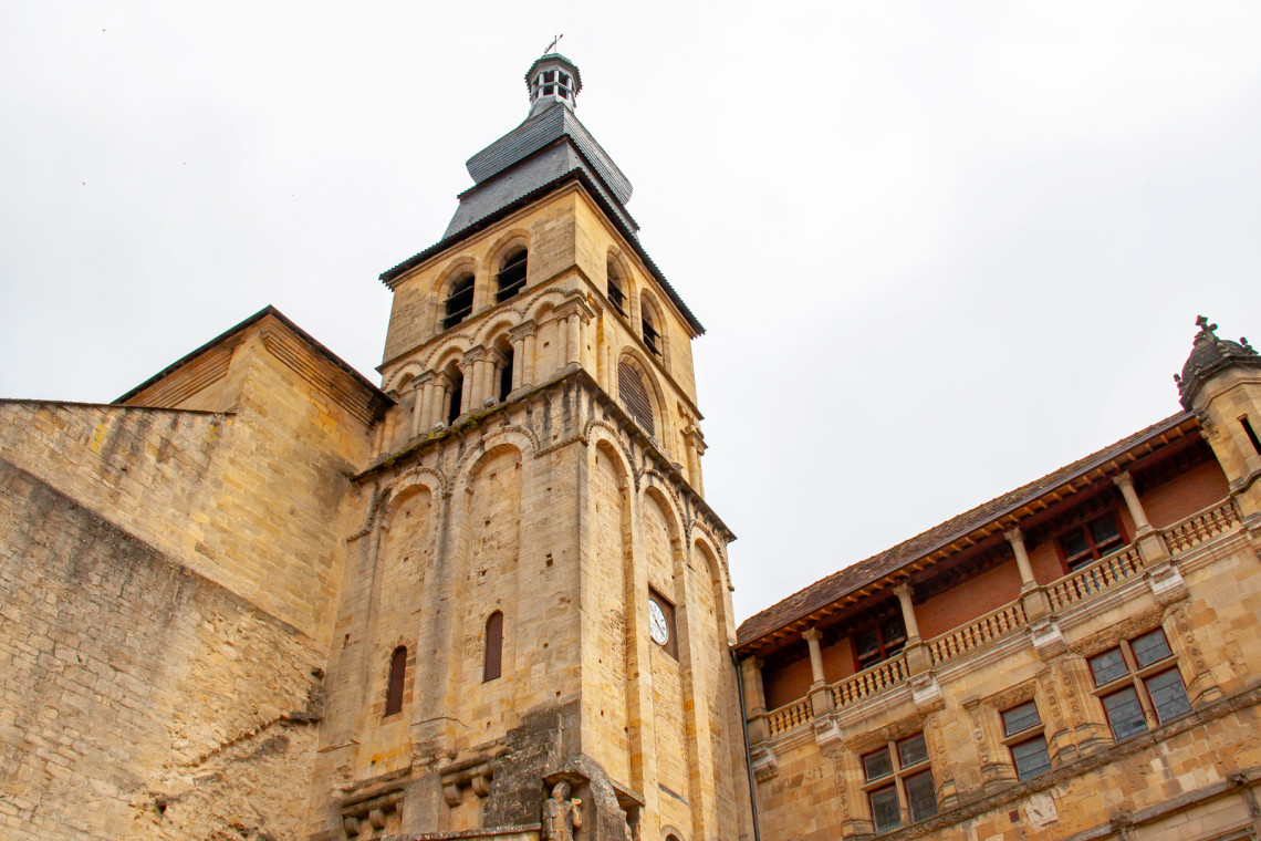  Low-angle view of the Romanesque bell tower of the former cathedral in Sarlat-la-Can&eacute;da, built in golden limestone with ornate arched belfry and clock face, with a Renaissance loggia building visible to the right