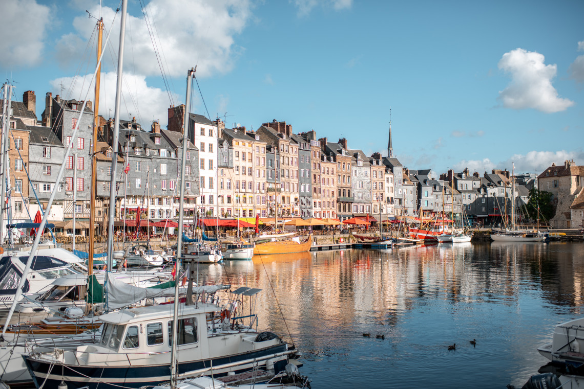 Honfleur's Vieux Bassin harbour in Normandy, with tall narrow slate-fronted and colourful painted townhouses lining the quay, red caf&eacute; awnings at street level, sailboats and fishing vessels moored in the foreground, and their reflections in the calm harbour water