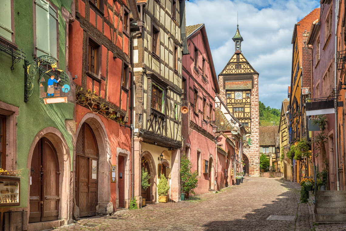 A cobblestone street in Riquewihr on the Alsace Wine Route, lined with intensely colourful half-timbered buildings in green, red and ochre, an ornate wrought-iron shop sign on the left, and the medieval Dolder gate tower visible at the end of the street