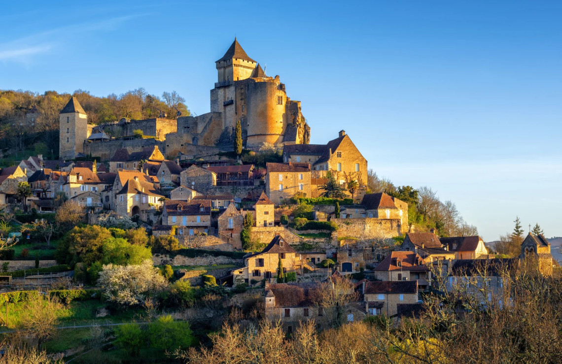 The medieval Ch&acirc;teau de Castelnaud crowning a hilltop village in the Dordogne, with golden limestone towers and ramparts above a cluster of stone houses cascading down the slope, surrounded by early spring woodland in warm afternoon light