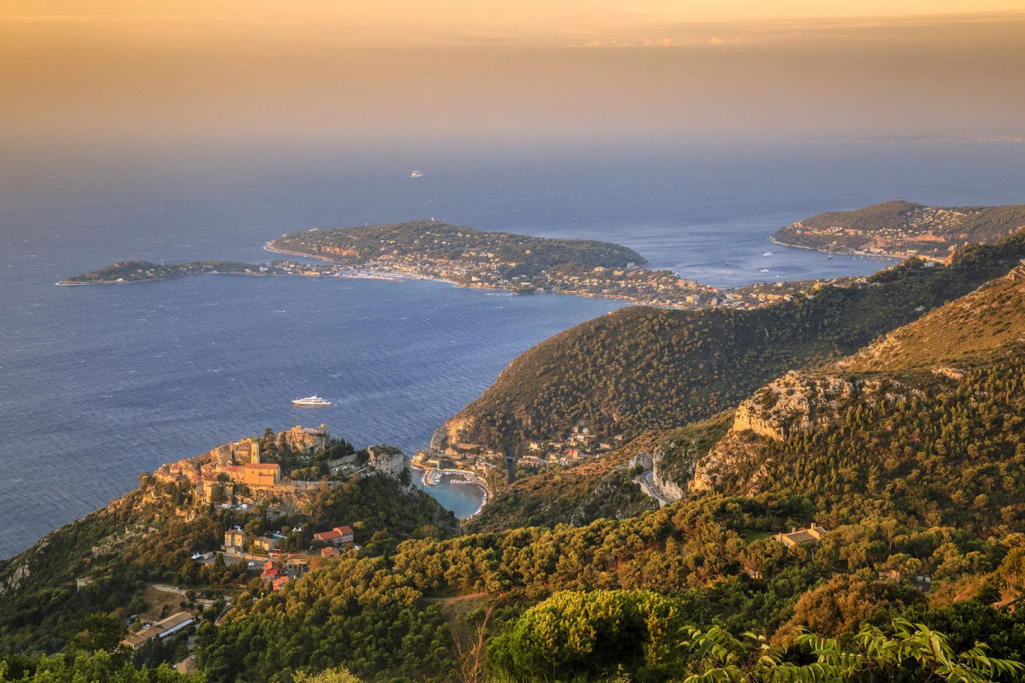 Aerial golden hour view from above &Egrave;ze on the French Riviera, showing the medieval village perched on a rocky clifftop promontory, with the Mediterranean coastline, the wooded peninsula of Cap Ferrat, and a cruise ship in the distance below an orange sunset sky