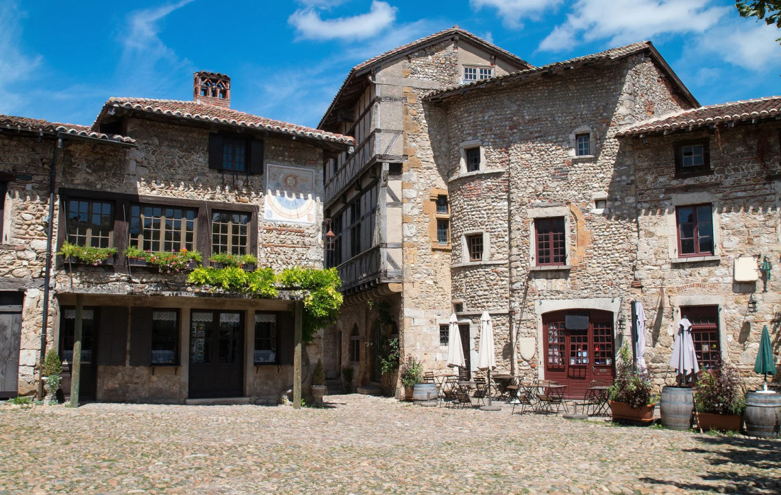 The main cobblestone square of the medieval walled village of P&eacute;rouges in Ain, with heavy stone and timber-framed buildings, an outdoor caf&eacute; terrace, wine barrel planters, and climbing vines on the facade under a blue summer sky