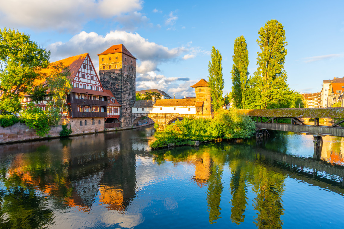 The medieval towers of the Ponts Couverts reflected in the calm River Ill in Strasbourg's Petite France quarter, with a half-timbered building on the left, a covered wooden bridge on the right, and tall poplar trees in warm evening light