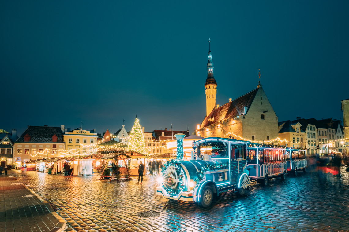 Tallinn's Town Hall Square Christmas market at night with a blue LED-lit tourist train in the foreground, illuminated market stalls, a large Christmas tree, and the Gothic Town Hall tower rising behind under a deep blue sky
