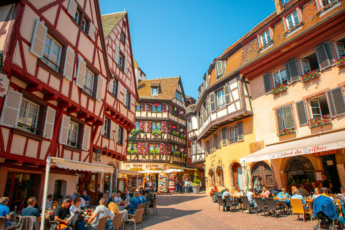 Place de l'Ancienne Douane in Colmar on a sunny summer day, with busy outdoor caf&eacute; terraces on both sides, colourful half-timbered buildings enclosing the square, and the Bistrot du Ko&iuml;fhus sign visible on the right