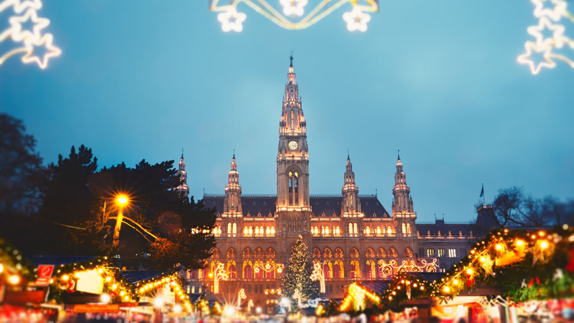 The illuminated Neo-Gothic Vienna Rathaus at dusk during the Wiener Christkindlmarkt, with rows of glowing Christmas market stalls and a large Christmas tree in the foreground, and star-shaped light decorations framing the top of the image