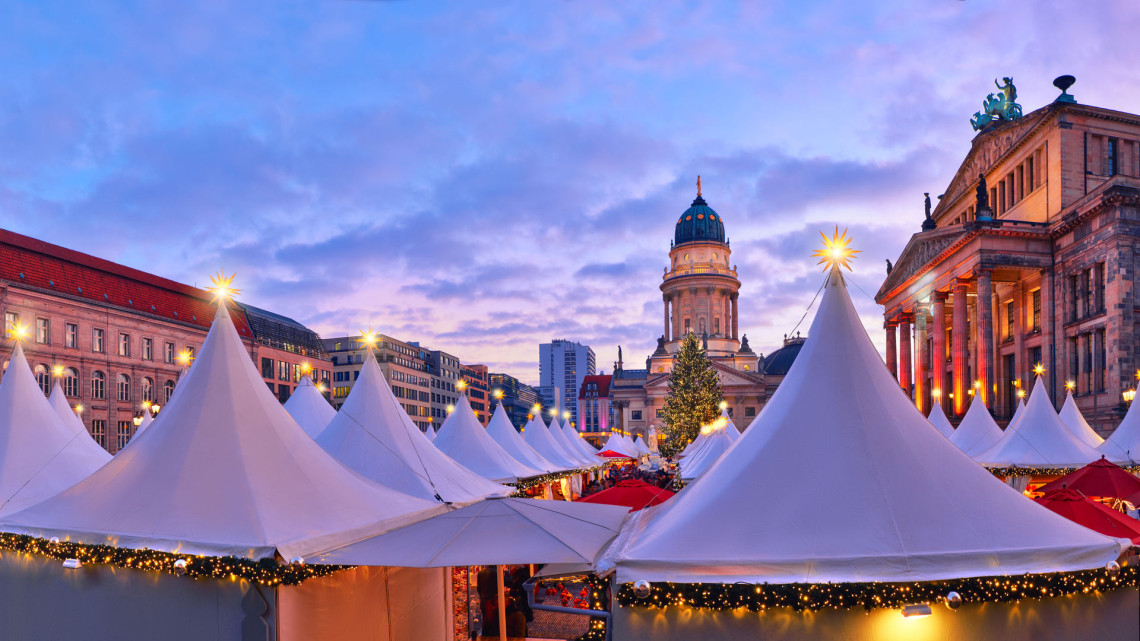 The Gendarmenmarkt Christmas market in Berlin at dusk, with rows of white peaked market tents decorated with fairy lights in the foreground, the green-domed Franz&ouml;sischer Dom in the centre, and the illuminated Konzerthaus colonnade on the right under a purple evening sky