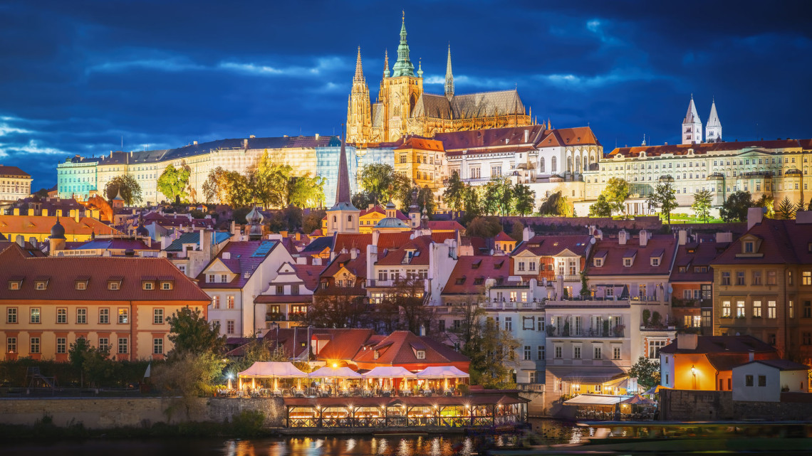 Prague Castle and the illuminated Gothic spires of St. Vitus Cathedral at twilight, rising above the red-roofed buildings of Mal&aacute; Strana, with the Vltava riverbank and a lit restaurant terrace in the foreground under a deep blue sky