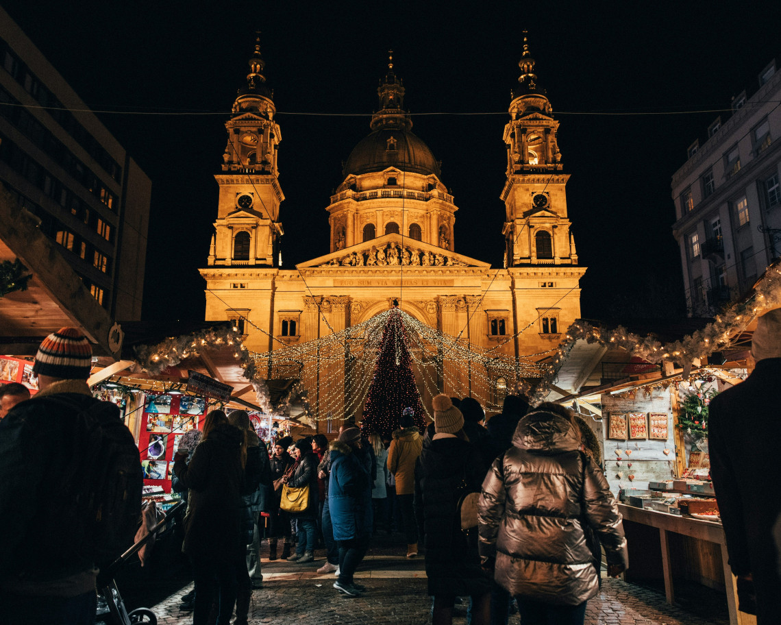 The Christmas market in front of St. Stephen's Basilica in Budapest at night, with crowds browsing market stalls, a large lit Christmas tree framed by the illuminated Neoclassical basilica facade and its twin towers in the background
