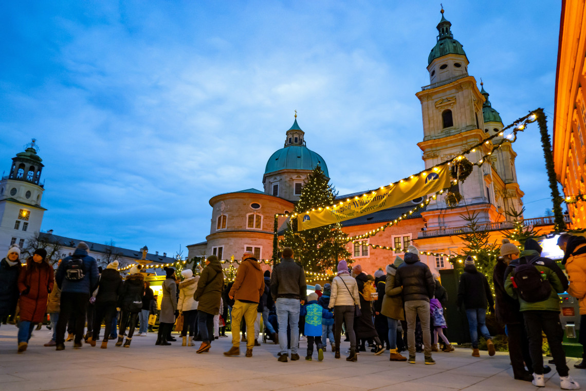 Crowds at the Salzburg Christmas market at dusk, with a large Christmas tree, fairy-lit market stalls, and the green copper dome and towers of a Baroque cathedral rising behind under a blue evening sky