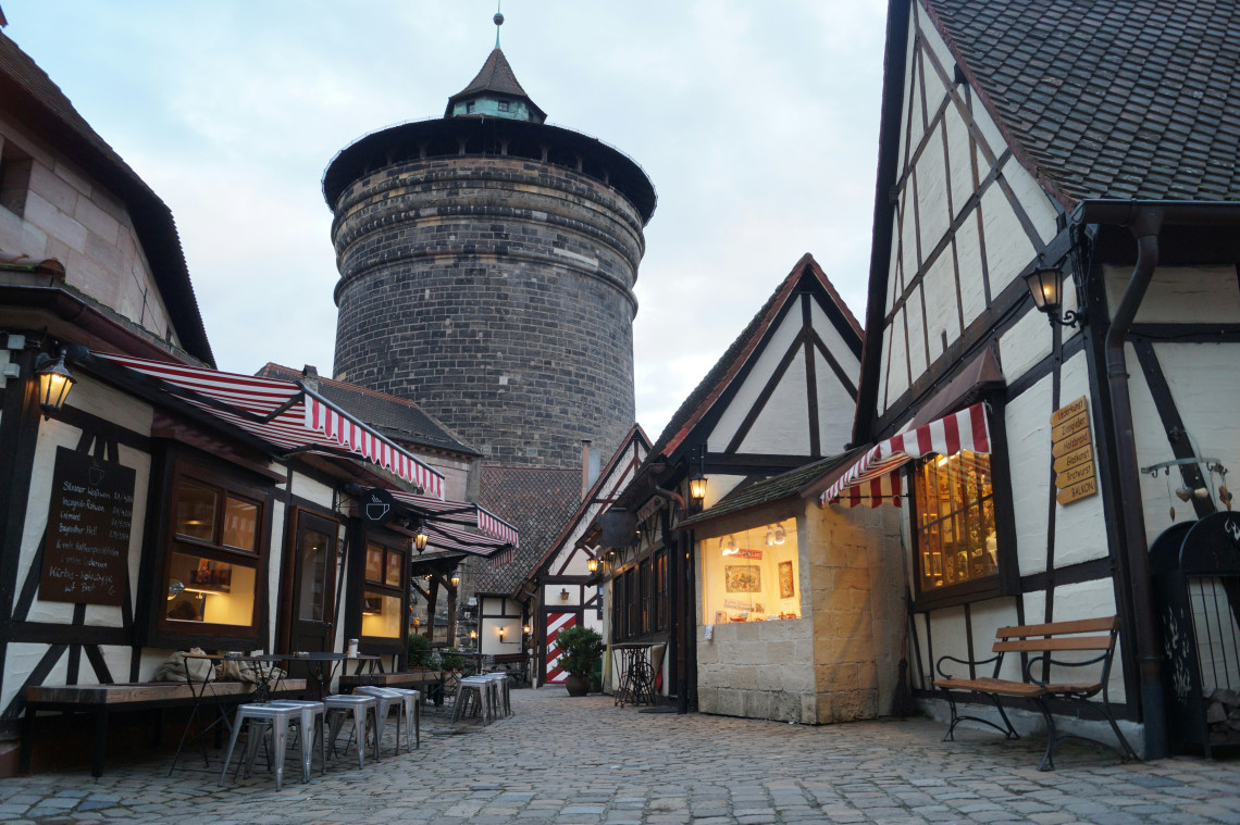 The Handwerkerhof artisan courtyard in Nuremberg at dusk, with half-timbered craft shops and red-and-white striped awnings surrounding a cobblestone square, and the large round dark stone Neutorturm city wall tower rising behind