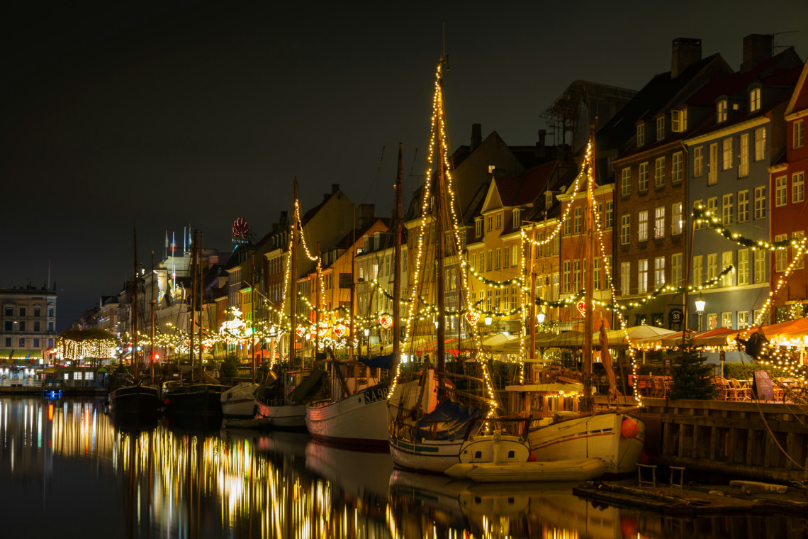 Copenhagen's Nyhavn canal at night during Christmas, with historic wooden sailing boats with masts wrapped in fairy lights moored along the quay, colourful 17th-century townhouses reflected in the still dark water, and Christmas market stalls visible along the right bank