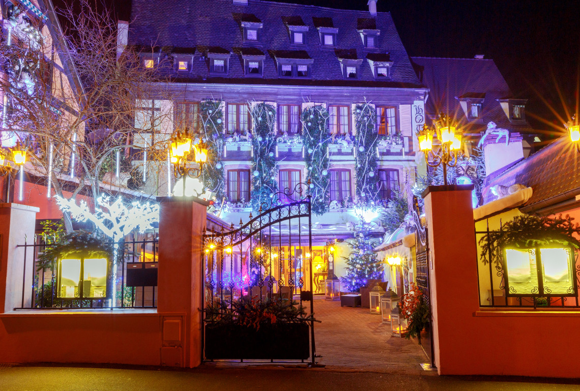 A grand French mansion at night decorated for Christmas, with ornate wrought-iron entrance gates, trees wrapped in white and blue LED lights, a purple light wash on the facade, and warm street lanterns on either side