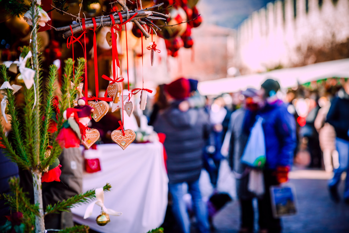 Close-up of a Christmas market stall with gingerbread heart ornaments and red-ribboned decorations hanging in the foreground, a small Christmas tree branch, and a crowd of shoppers blurred in the background