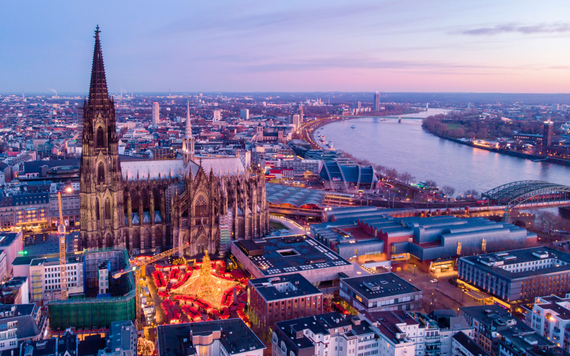 Aerial evening view of Cologne Cathedral and its illuminated Christmas market in Cologne, Germany. The Rhine River and Hohenzollern Bridge extend through the city in the background beneath a pastel winter sky.