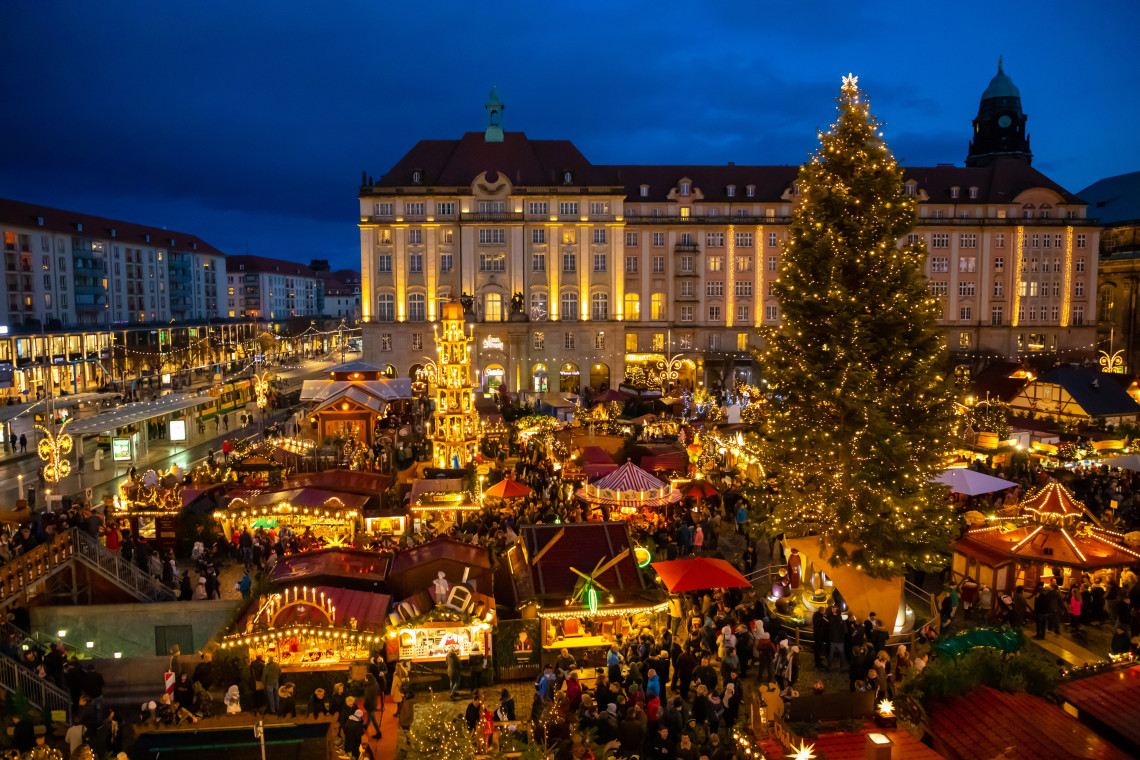 Night view of the Dresden Striezelmarkt Christmas market in Dresden, Germany, filled with illuminated wooden stalls and visitors. A large decorated Christmas tree rises above the market against the historic buildings of the city centre.